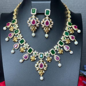 May include: A gold-toned necklace and earring set featuring rectangular green stones, teardrop-shaped pink stones, and clear white crystals. The jewelry is accented with small white pearls hanging from the bottom edges of the necklace and earrings.