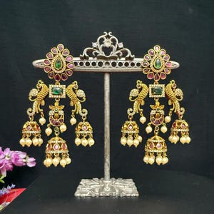 May include: Gold-toned earrings with intricate detailing. Each earring features a floral top with a green center stone and surrounding red gems. Below, a peacock design leads to a cluster of pearls and bell-shaped dangles. The earrings are displayed on a silver stand.