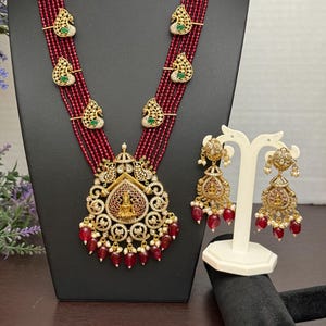 May include: A maroon beaded necklace and matching earrings set. The necklace features a large pendant with intricate gold detailing, clear and green gemstones, and dangling red beads. The earrings have a similar design, with gold accents and red bead drops.