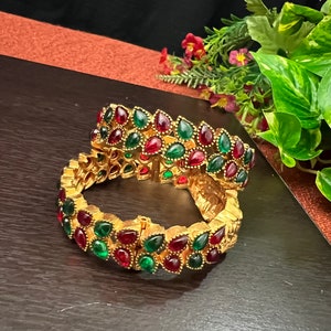May include: Two gold-colored bangles with alternating red and green teardrop-shaped gemstones. The bangles are displayed on a dark brown surface, with greenery and flowers in the background.