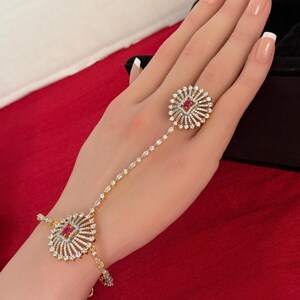 May include: A gold-toned hand chain bracelet with a ring and a bracelet element. The ring and bracelet feature a central red stone surrounded by a sunburst design of clear stones. The chain connecting the ring and bracelet is adorned with small, clear stones.