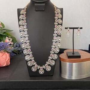 May include: A long gold-tone necklace featuring a series of circular pendants encrusted with clear white stones and accented with small red stones, paired with matching drop earrings that mirror the circular design and stone arrangement.