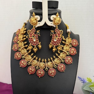 May include: A gold-toned jewelry set featuring a necklace and matching earrings. The necklace and earrings have intricate designs with red and green accents. The earrings are peacock-shaped, and the necklace has a series of circular pendants.