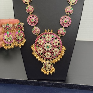 May include: A gold-toned necklace and matching earrings set. The necklace features a large, circular pendant with a floral design, surrounded by red and white stones. The earrings mirror the design. Green and pink stones are also incorporated.