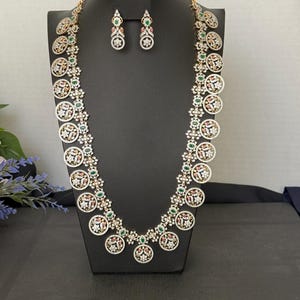 May include: A long gold-tone necklace featuring a series of circular pendants adorned with clear, green, and pink stones, paired with matching drop earrings. The jewelry set displays intricate floral patterns and pavé-style stone settings.