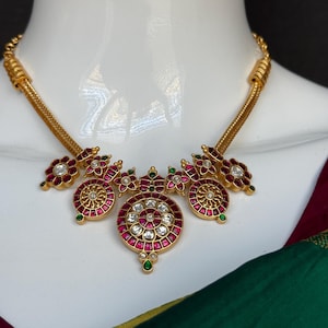 May include: A gold-toned necklace featuring a central circular pendant with embedded white stones and red accents. The design includes smaller floral-shaped motifs with red and green stones, connected by a textured gold chain that rests on a white display.