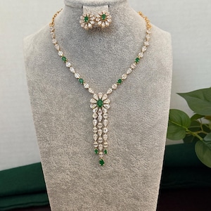 May include: A gold-toned necklace and matching stud earrings set featuring clear pear-shaped and round-cut stones arranged in a floral pattern. The necklace includes a central pendant with three dangling strands and small green oval-cut stones.