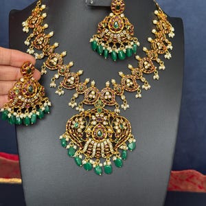 May include: A gold-toned necklace and earring set with intricate detailing. The jewelry features red and green gemstones, small white pearls, and dangling green beads. The set is displayed on a black mannequin against a dark blue backdrop.