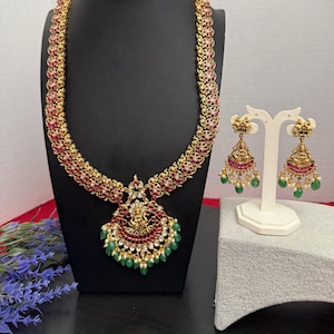 May include: A gold-toned necklace and earring set. The necklace features a pendant with a central figure, red and green accents, and a series of green beads. The earrings mirror the design with red and green details.