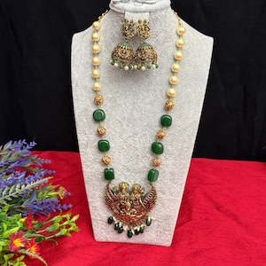 May include: An ornate necklace and earring set displayed on a mannequin. The necklace features a pendant with figures, green square beads, and faux pearls. The earrings have a matching design. The jewelry is gold-toned with green and cream accents.