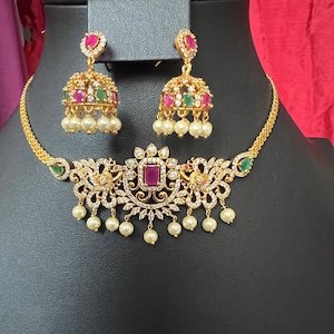 May include: Gold-toned jewelry set featuring a necklace and matching earrings. The necklace has a central square ruby-colored stone, surrounded by clear crystals and pearl drops. The earrings have ruby and emerald-colored stones and pearl drops.