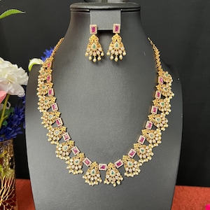 May include: A gold necklace with pink and green stones and a matching pair of earrings. The necklace has a pendant design with pearl accents.