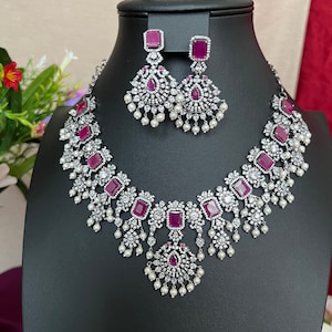 May include: A silver necklace and earrings set with a pink stone and white cubic zirconia accents. The necklace features a layered design with dangling pearls.