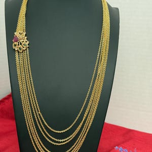 May include: A gold-colored necklace with multiple strands of delicate chains. The necklace features a decorative floral pendant with red and green gemstones. The necklace is displayed on a black mannequin.