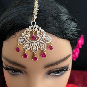 May include: A gold-tone forehead chain with a teardrop-shaped pendant adorned with clear cubic zirconia stones and pink gemstones. The chain is decorated with a row of clear cubic zirconia stones.