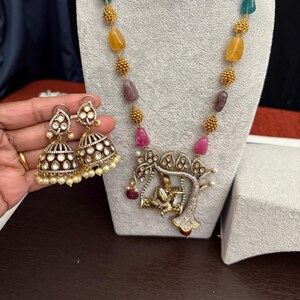 May include: A gold-toned necklace and matching earrings set. The necklace features a pendant with a figure under a tree, and colorful gemstone beads. The earrings are bell-shaped with white stones and pearls.