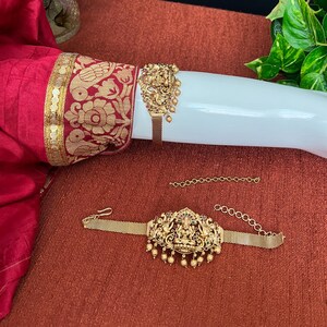 May include: A gold-toned metal bracelet with a detailed design featuring a deity figure, green and red gemstones, and dangling beads. The bracelet is on a white mannequin arm.