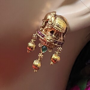 May include: Gold elephant-shaped earrings with dangling green and red gemstones. The earrings have a detailed design and are a traditional Indian style.