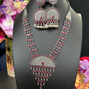 Puede incluir: Un collar de plata con un colgante que presenta una piedra roja y una franja de piedras rojas. El collar está formado por tres hileras de piedras rojas. También se incluyen pendientes a juego con piedras rojas.