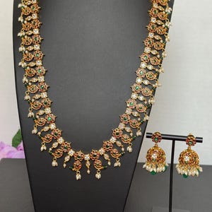 May include: A gold-toned necklace and matching earrings set. The necklace features a repeating pattern of red, green, and clear gemstones, with small white pearls. The earrings are bell-shaped with similar detailing. The jewelry is displayed on a black stand.