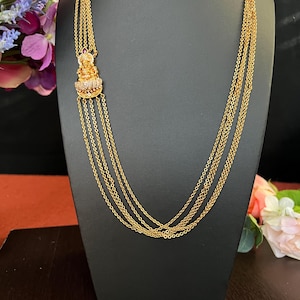 May include: A gold layered necklace with a small pendant featuring a detailed design. The necklace is displayed on a black stand.