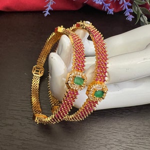 May include: Two gold bangles with a pink and white stone design. The bangles are decorated with emerald green stones.