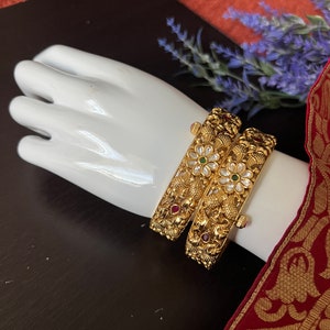 May include: Two gold bangles with intricate floral designs and embedded gemstones. The bangles are adorned with white, red, and green stones.