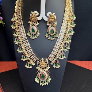 May include: A gold necklace set with a large pendant featuring a green and red stone. The necklace is adorned with white stones and green beads. The earrings are similar in design to the pendant.
