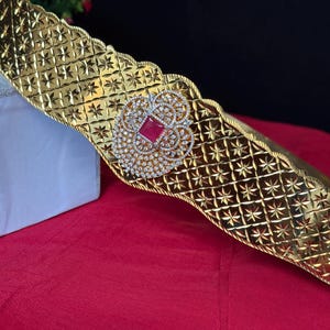 May include: A gold-colored belt with a repeating starburst pattern. The belt features a central, ornate embellishment with a red square gemstone surrounded by clear, sparkling stones. The belt's edges have a scalloped design.