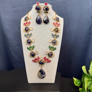 May include: A gold-toned necklace and earring set. The necklace features a series of dark blue, red, and green gemstones. The earrings have a similar design, with a dark blue teardrop-shaped stone. The jewelry is displayed on a white mannequin.