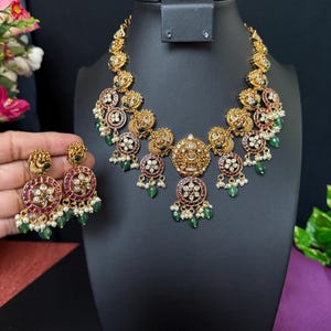 May include: A gold-toned necklace and matching earrings set. The necklace and earrings feature intricate floral designs with red, green, and white accents. The set is displayed on a black jewelry stand.