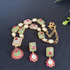 May include: A gold-toned necklace and earring set. The necklace features square and rectangular green and pink stones with red and clear accents. The earrings have matching green stones and red and clear accents. The jewelry is set against a black background.
