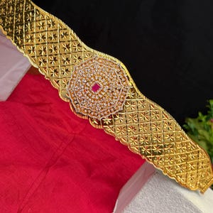 May include: A gold-colored waist belt with a central, circular design encrusted with small, clear stones and a central red stone. The belt has a repeating starburst pattern and a textured surface.