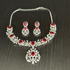 May include: A silver necklace and earrings set with a red stone pendant and red stone earrings. The necklace has a floral design with small clear stones.