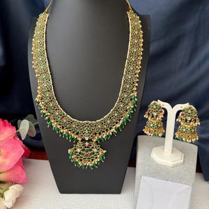 May include: A gold necklace with a green and white floral design and a matching pair of earrings. The necklace has a pendant in the shape of a bell.