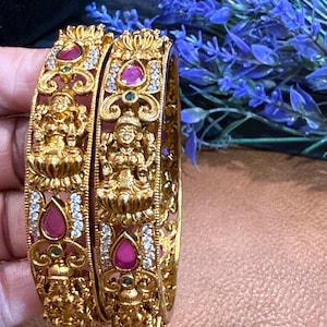 May include: Two gold bangles featuring intricate embossed figures of a deity seated on a lotus flower. The metalwork is accented with teardrop-shaped pink stones, small clear crystals, and tiny green stones set within ornate filigree patterns.