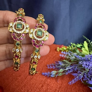 May include: Two gold-toned bangles with intricate floral designs. Each bangle features a central square green stone surrounded by a circle of white stones and smaller red stones. The bangles are adorned with additional green and red stones.