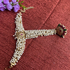 May include: A gold and pearl headpiece with a red and green gemstone design. The headpiece is made of a chain with multiple strands of pearls.
