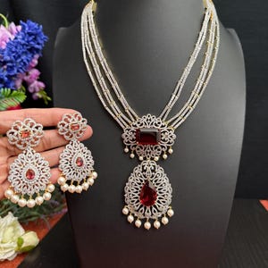 May include: A multi-strand necklace with a large red gemstone pendant and matching earrings. The necklace and earrings are adorned with clear cubic zirconia stones and small white pearls.