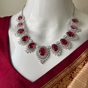 May include: A silver necklace with a row of oval-shaped red gemstones surrounded by smaller clear gemstones. The necklace is on a white mannequin bust against a maroon and gold background.