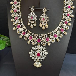 May include: A necklace and earring set featuring silver-toned metal filigree adorned with rectangular and teardrop-shaped fuchsia stones, clear pavé crystals, and small white pearls. The necklace includes a large central floral-shaped pendant.