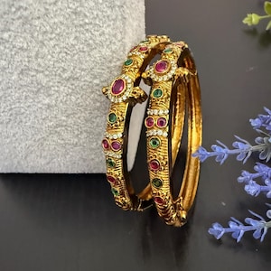 May include: Two gold bangles with intricate detailing, adorned with red, green, and clear gemstones. The bangles have a textured surface and are set against a gray and black background. The gemstones are round and oval-shaped.