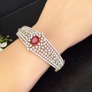 May include: A gold-toned bracelet with a central oval ruby and numerous small, clear gemstones. The bracelet is set with a pattern of sparkling stones, creating a luxurious and elegant appearance. The clasp is also gold-toned.