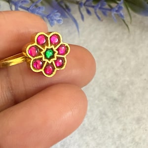 May include: A gold-colored ring with a flower-shaped design. The flower has pink and green gemstones. The ring is held by a person's fingers.