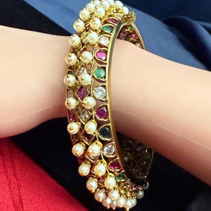 May include: A gold-toned bangle bracelet adorned with pearls and colorful gemstones. The bracelet features a series of alternating ruby, emerald, and clear stones, with a row of pearls along the top. The bracelet is worn on a wrist.