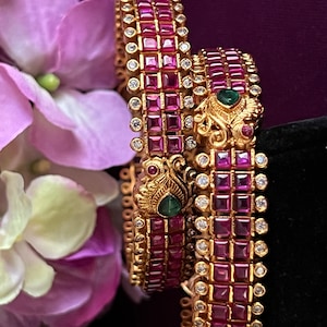 May include: Two gold bangles with pink and green stones. The bangles are decorated with intricate designs and are set with small, square-shaped stones.