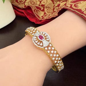 May include: A gold-toned hinged bracelet featuring a central circular ornament set with a single oval-shaped red gemstone surrounded by small clear stones. The band is decorated with multiple rows of small clear stones in a grid-like pattern.
