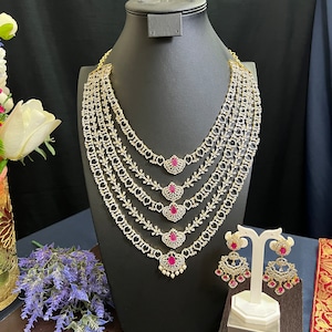 May include: A five-strand gold necklace with white stones and pink accents. The necklace is displayed on a black mannequin. Matching earrings with pink stones are displayed on a white stand.