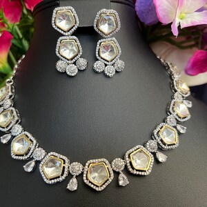 May include: A silver and gold necklace with a row of large, clear, faceted stones set in a geometric pattern. The necklace is accented with smaller clear stones and has a matching pair of earrings with a similar design.