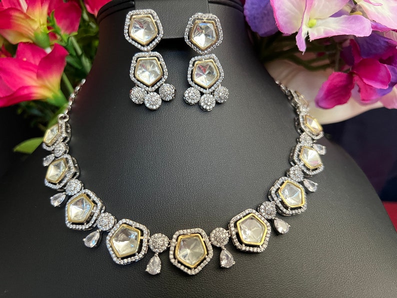 May include: A silver and gold necklace with a row of large, clear, faceted stones set in a geometric pattern. The necklace is accented with smaller clear stones and has a matching pair of earrings with a similar design.